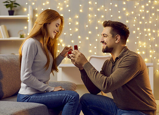 Couple engaged in a romantic proposal with the man holding an engagement ring box in front of the woman, surrounded by soft glowing fairy lights.