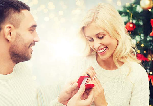 Man proposing to his girlfriend with an engagement ring box during Christmas, with a joyful woman smiling and admiring the ring in front of a festive tree.