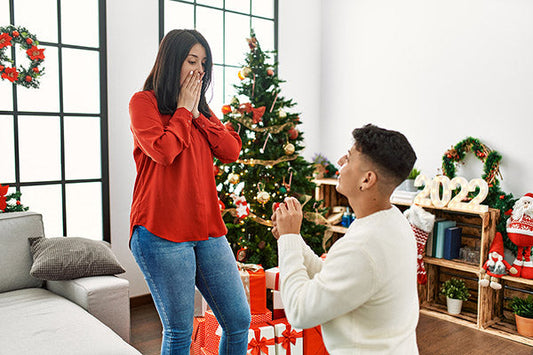 Man proposing to his girlfriend during Christmas season in front of a decorated tree with presents around, woman surprised and covering her face in joy.