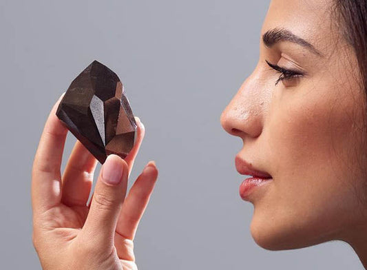 Close-up of a woman holding a large rough black diamond, admiring its facets. She is gently examining the gemstone, highlighting its natural structure and brilliance, set against a neutral gray background.