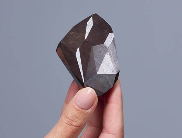 Close-up of a person holding a large rough black diamond with distinct facets. The diamond is shown in hand, highlighting its natural structure and impressive size, set against a neutral gray background.