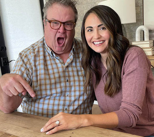 A man and a woman smiling and sitting at a table, with the man pointing excitedly at a diamond engagement ring on the woman's hand, showcasing their joyful moment.