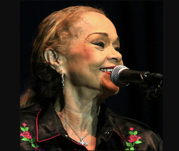 Etta James smiling while singing into a microphone on stage, wearing a black shirt with red rose embroidery and earrings, with soft lighting on her face.