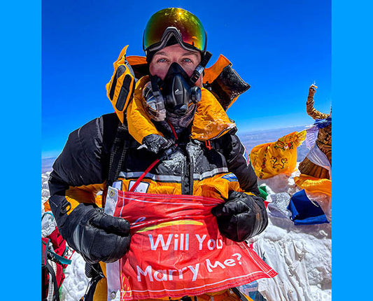 Person in mountaineering gear holding a 'Will You Marry Me?' flag at the summit of Mount Everest, with the clear blue sky and colorful prayer flags in the background.
