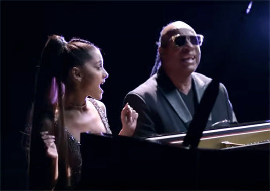 Ariana Grande and Stevie Wonder performing together at a piano, both smiling and enjoying the performance, with Ariana wearing a sparkling outfit and Stevie wearing sunglasses.