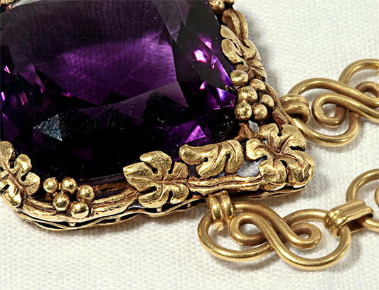 Close-up of a vintage gold pendant featuring a large purple amethyst gemstone, surrounded by intricate floral designs. The pendant's detailed craftsmanship is highlighted, showcasing the elegant goldwork and the vibrant gemstone.