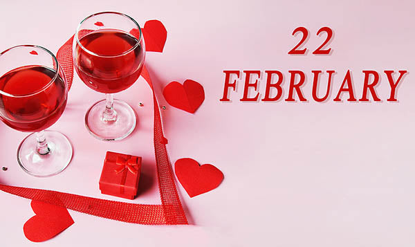Valentine's Day themed image featuring two glasses of red wine, a small red gift box, and red heart-shaped decorations. The date '22 FEBRUARY' is prominently displayed against a soft pink background, evoking a romantic atmosphere.