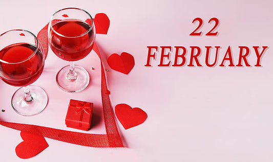 Valentine's Day themed image featuring two glasses of red wine, a small red gift box, and red heart-shaped decorations. The date '22 FEBRUARY' is prominently displayed against a soft pink background, evoking a romantic atmosphere.