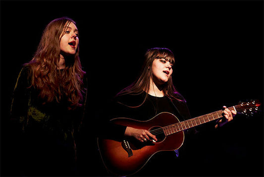 Two female musicians performing on stage, one singing and the other playing an acoustic guitar. The spotlight highlights their expressions as they sing together in a dark setting.