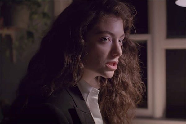Close-up of a woman with curly hair wearing a black blazer, looking thoughtfully to the side. The lighting creates a soft, moody atmosphere, with windows and soft shadows in the background.