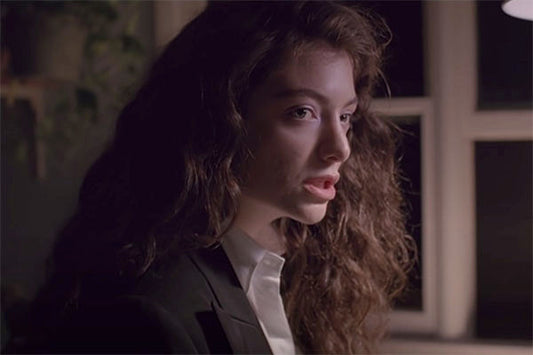 Close-up of a woman with curly hair wearing a black blazer, looking thoughtfully to the side. The lighting creates a soft, moody atmosphere, with windows and soft shadows in the background.