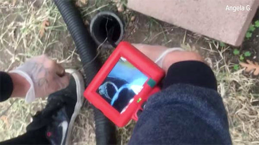 Person using a pipe inspection camera to check a drainage pipe, displaying an image of a heart-shaped object inside the pipe on a small monitor screen.