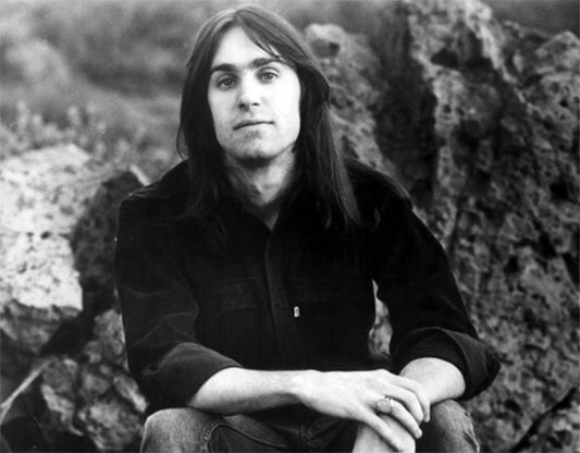 Black and white photo of a young man with long hair sitting on rocks, wearing a dark shirt, and looking at the camera.