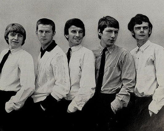 A black-and-white photograph of five men posing in a line, dressed in formal attire with button-up shirts and ties. They stand side by side with their hands in their pockets, offering a classic 1960s or 1970s style look.