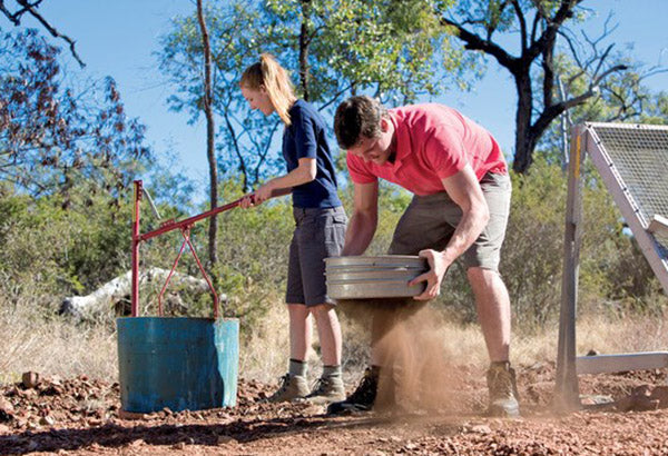 Two people panning for gold in the Australian outback, with one person sifting dirt and the other using a manual water pump.
