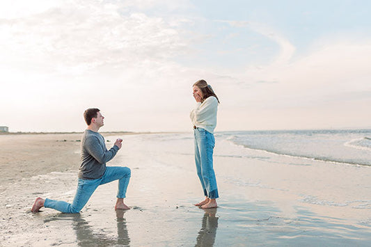 Man proposing to his partner on a beach, kneeling with an engagement ring, while the woman looks surprised and happy.