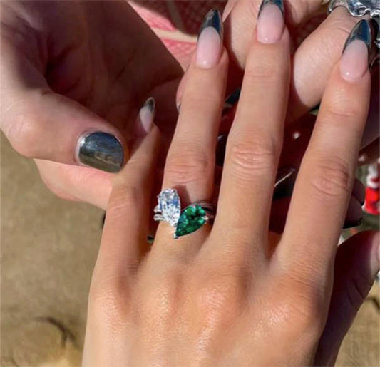 Close-up of a person's hand with an engagement ring featuring a large emerald pear-shaped stone and a smaller diamond. The ring is being admired as a hand is placed on the ring finger, with the focus on the stunning design and gemstones.
