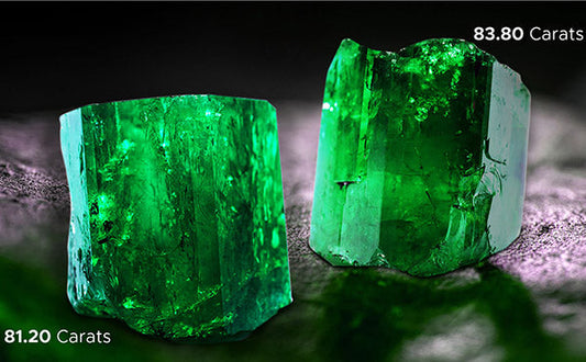 Close-up of two large emerald gemstones, one weighing 81.20 carats and the other 83.80 carats, with vibrant green color and intricate textures, placed side by side on a dark background.