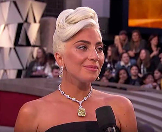 Lady Gaga at the Oscars wearing a glamorous diamond necklace and earrings, with a chic updo hairstyle and a strapless black dress, smiling during an interview on stage.