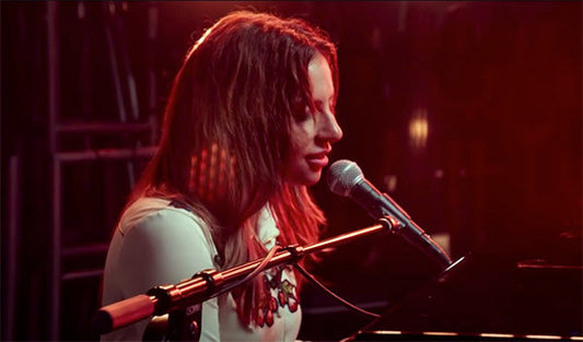 Lady Gaga performing at a piano, singing into a microphone with soft lighting in the background, capturing an intimate performance moment.