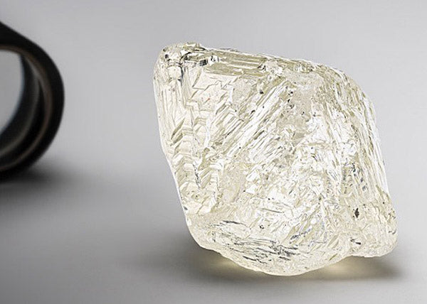 Large raw diamond with rough, faceted edges resting on a neutral surface, showing the unique texture and clarity of the natural gemstone before being cut and polished.