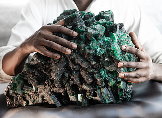 A person holding a large, rough emerald crystal cluster, showcasing vibrant green and dark mineral formations.