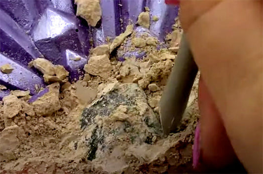 Close-up of a person using a tool to carefully uncover a gemstone from the surrounding dirt.