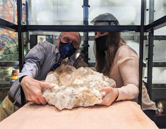 Two people wearing masks carefully handling a large, crystal-like mineral specimen inside a display case at a museum or exhibition.