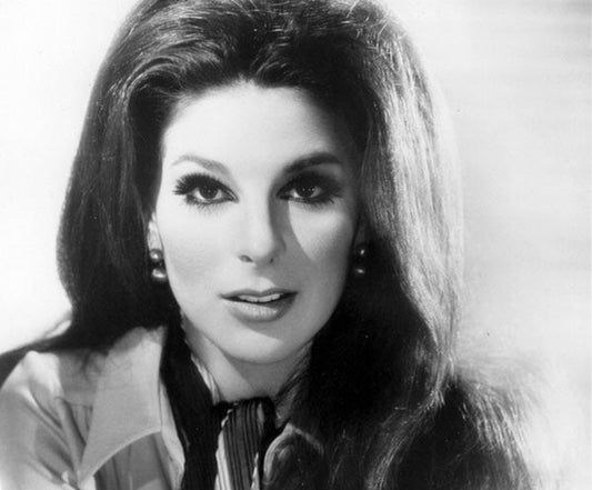 Black-and-white portrait of a woman with long, voluminous hair and bold makeup, including dramatic eyeliner. She is wearing pearl earrings and a blouse with a scarf, exuding a glamorous vintage look.