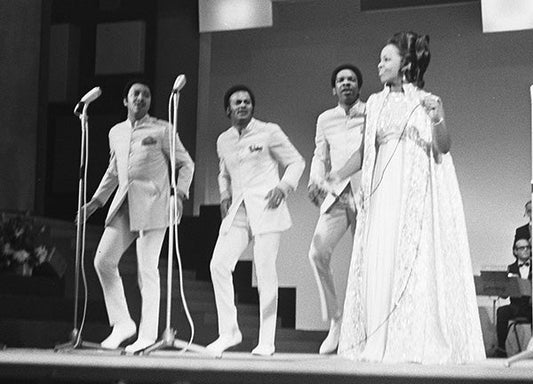 A vintage black-and-white scene of a musical performance on stage. A group of four performers, two men and two women, are dressed in formal attire, with the men wearing matching white suits and the woman in an elegant gown.