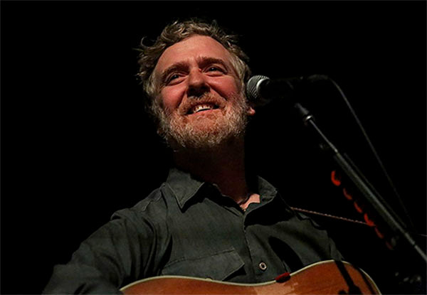 Portrait of musician Glen Hansard smiling while playing the guitar on stage, with a microphone in front of him.
