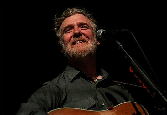 Portrait of musician Glen Hansard smiling while playing the guitar on stage, with a microphone in front of him.