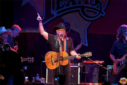 Willie Nelson performing on stage with his guitar during a live concert, raising his finger while other musicians play in the background, captured in a vibrant live music setting.