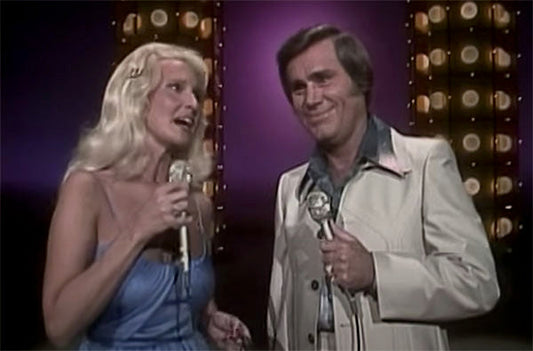 Classic image of a male and female singer performing a duet on stage with microphones, set against a backdrop of bright stage lights.