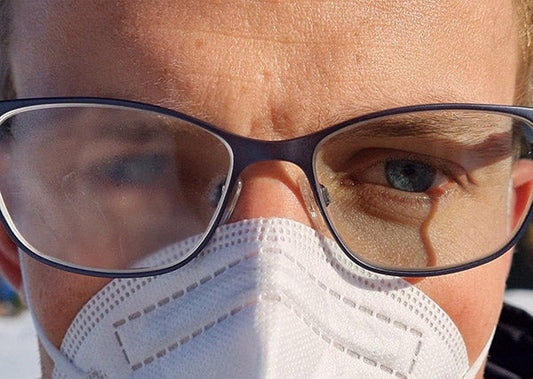 Close-up of a person wearing glasses with fogged lenses and a face mask, showing one eye clearly and the other slightly covered by fog.