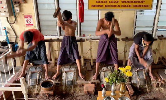 Four workers at a gold leaf workshop using hammers to shape metal into thin sheets, showcasing the traditional craftsmanship process in a workshop setting.