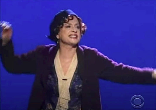 Actress performing on stage in a theatrical production, dressed in vintage 1920s clothing with a flapper-style hat and curly hair. She is joyfully singing or dancing with her arms outstretched, set against a blue background.