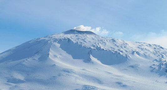 Snow-covered mountain peak with volcanic activity visible at the summit against a clear blue sky.