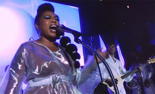 Singer performing on stage with a microphone, wearing a silver reflective outfit, accompanied by a musician playing the bass guitar, with colorful lighting in the background.