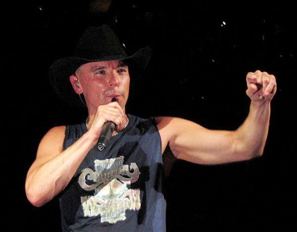 Kenny Chesney performing on stage in a cowboy hat and sleeveless shirt, holding a microphone and pointing to the audience.