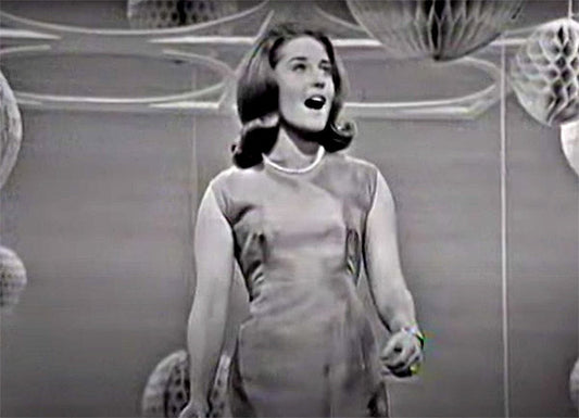 Black and white image of a woman singing on stage, wearing a dress and a pearl necklace, with paper lanterns in the background.