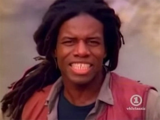 Smiling man with dreadlocks wearing a red vest, captured in a music video, flashing a bright smile.
