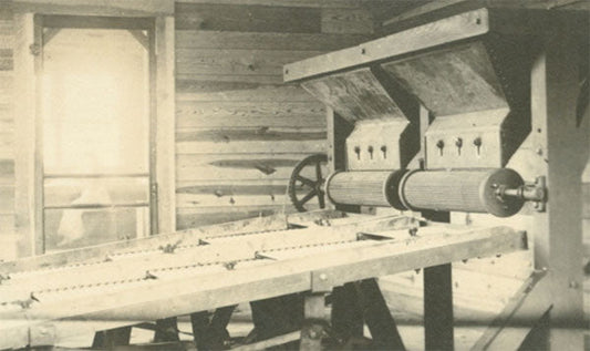 Historical black-and-white photo of a vintage mechanical machine with rotating cylinders, set inside a wooden building.