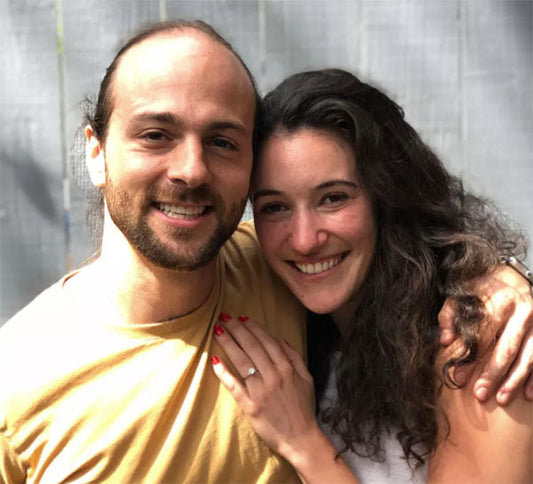 A happy couple smiling at the camera while embracing each other, with the woman showing her engagement ring on her left hand.