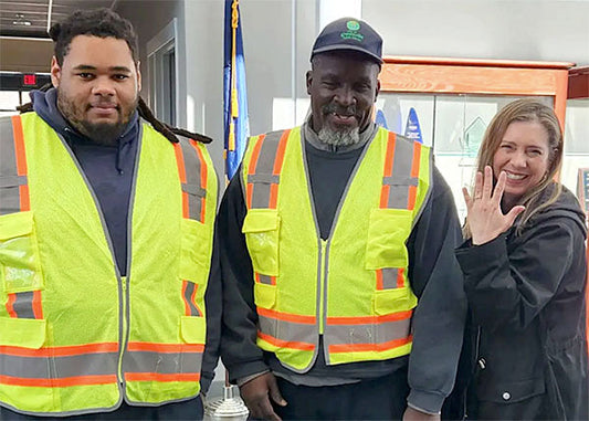Three individuals, two men and one woman, standing together in a professional setting. They are dressed in high-visibility safety vests, with the men wearing caps and the woman smiling and waving.