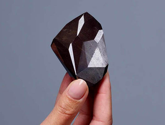Hand holding a large rough diamond with geometric facets and dark color.