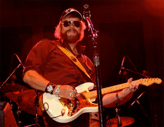 Hank Williams Jr. performing live on stage, playing an electric guitar with sunglasses and a cap. He is surrounded by band equipment, with a dynamic lighting setup creating a vibrant concert atmosphere.