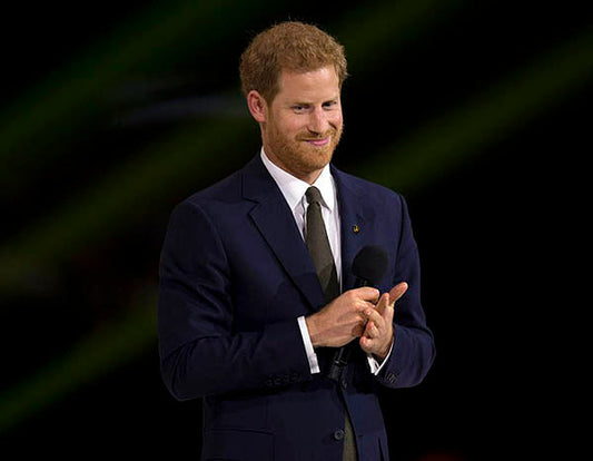Prince Harry smiling while holding a microphone, wearing a dark suit and tie, in a public speaking setting.