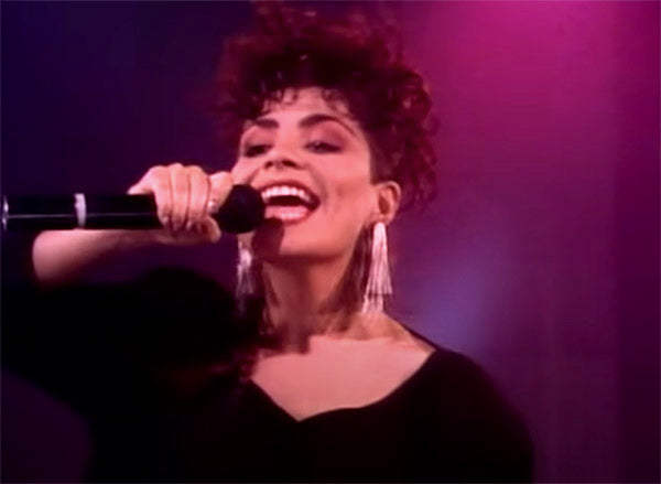 Female singer with curly hair performing on stage, holding a microphone and smiling, wearing dangling earrings, with vibrant pink lighting in the background.
