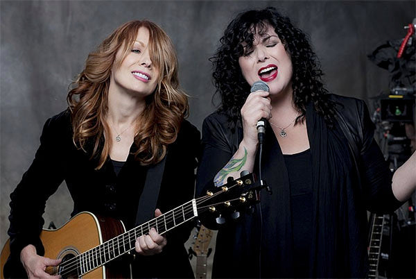 Heart duo performing, with one member playing guitar and the other singing into a microphone, both dressed in black outfits.